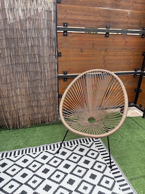 A wicker chair with a circular frame, positioned on a patterned rug, against a backdrop of a wooden wall with metal brackets and a bamboo fence.