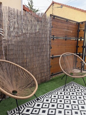 A fenced outdoor area with two wicker chairs and a patterned rug on artificial grass.