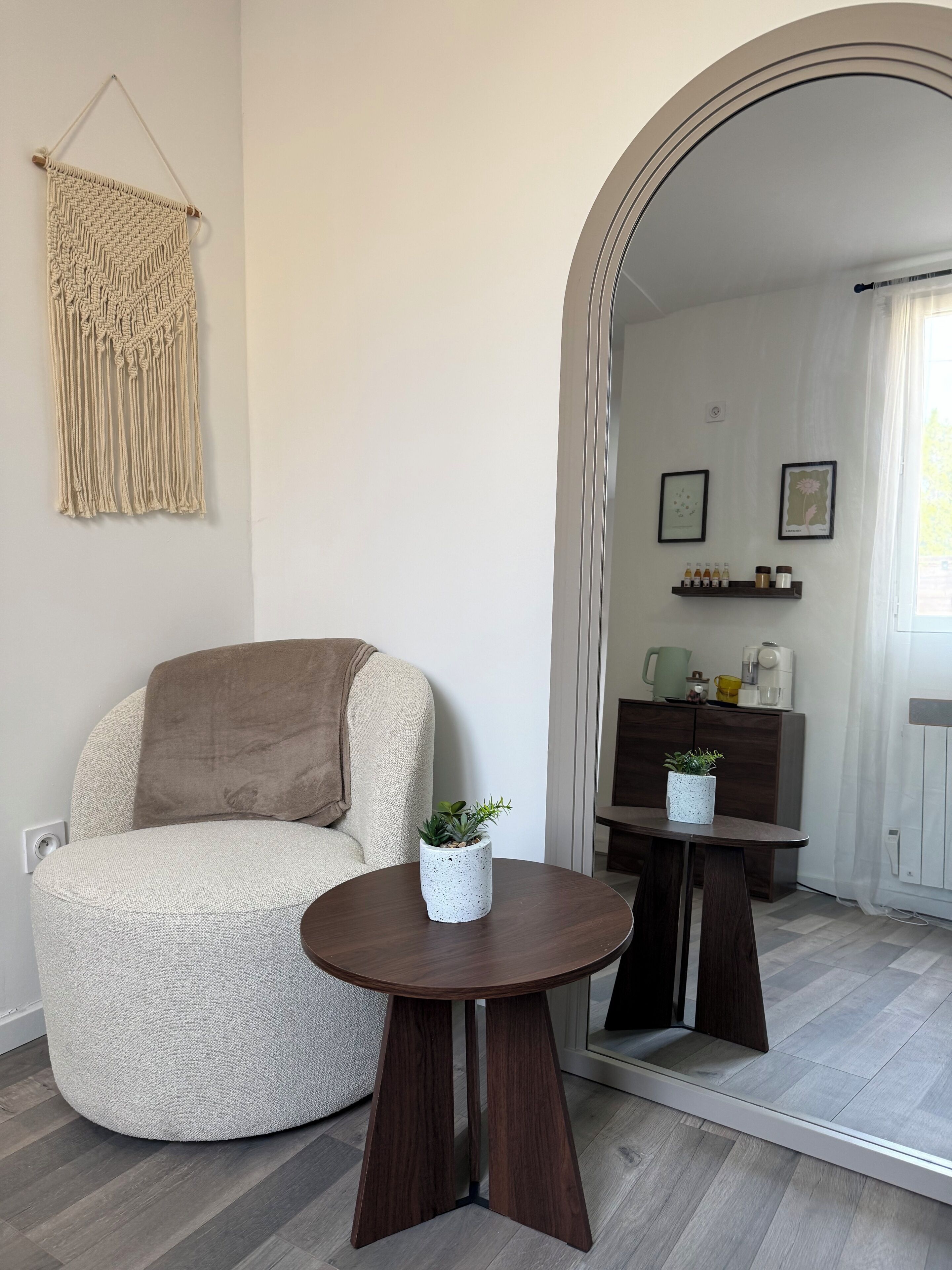 A modern living room with a round wooden table, a beige armchair, a macrame wall hanging, and a small potted plant.