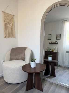 A modern living room with a round wooden table, a beige armchair, a macrame wall hanging, and a small potted plant.