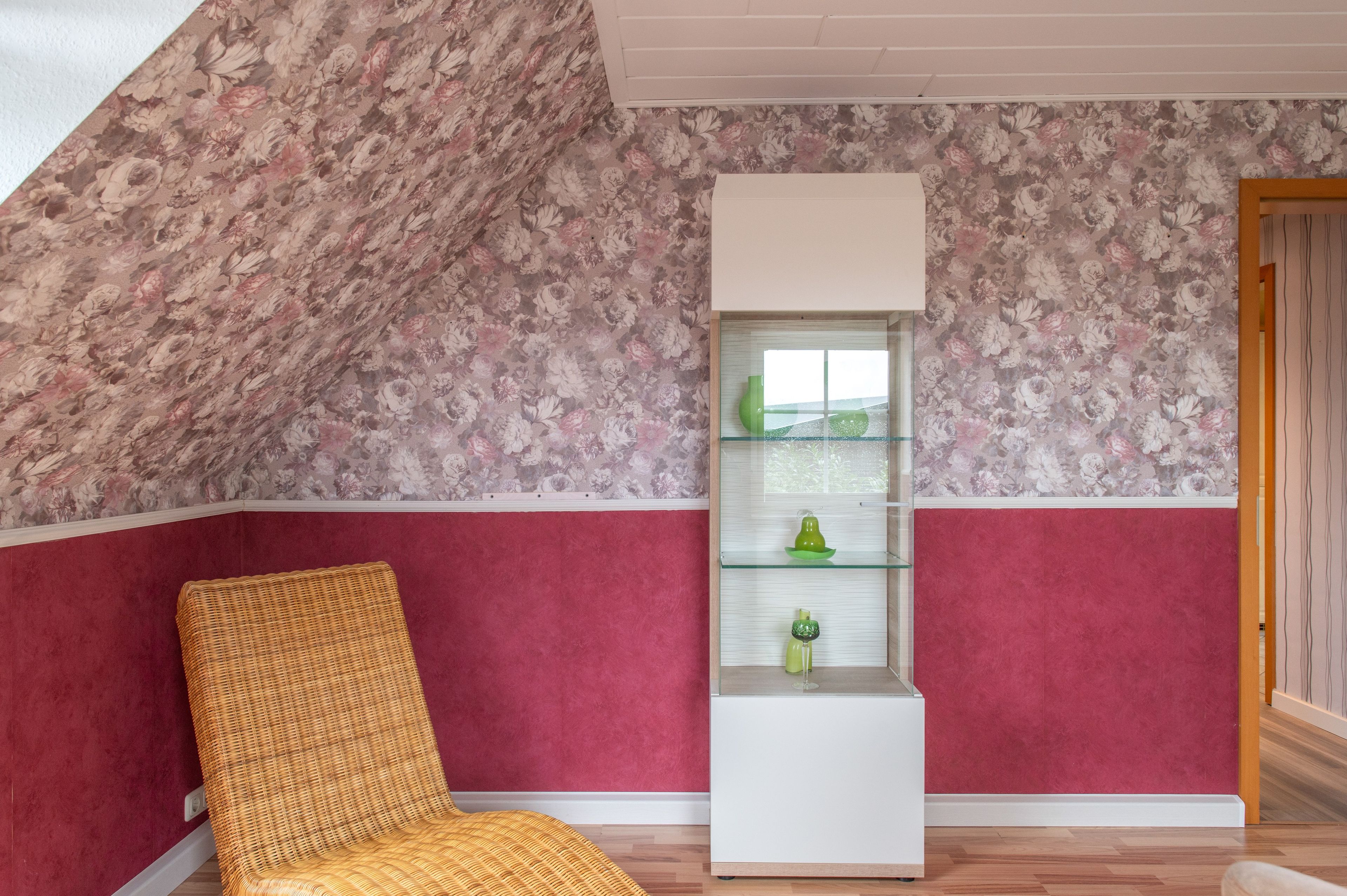 Een kamer met een schuine kap, een rieten stoel, een wit wandrek met glazen deuren en een rode en witte muur.
