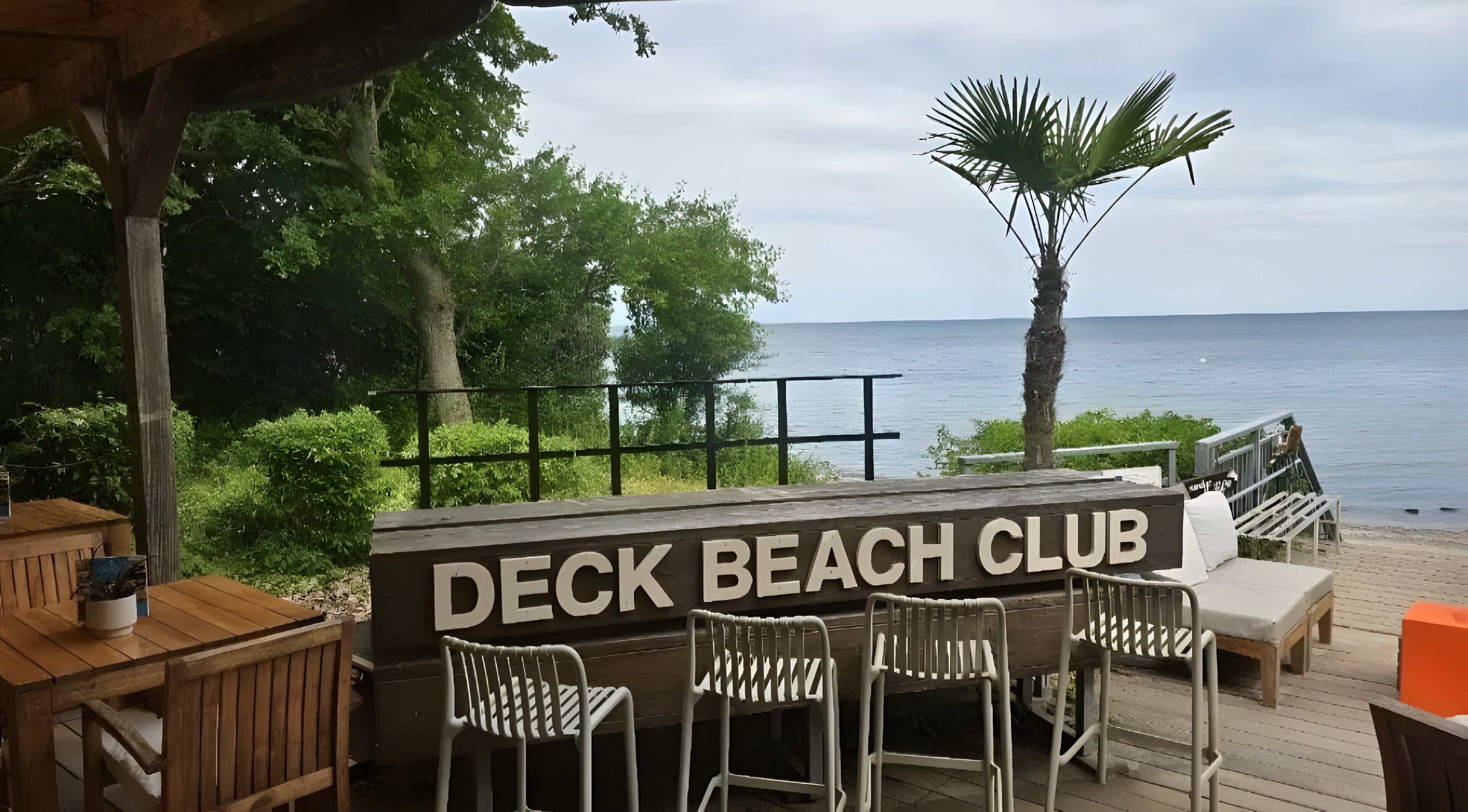 A wooden deck with a sign reading 'DECK BEACH CLUB,' surrounded by greenery and overlooking a body of water.
