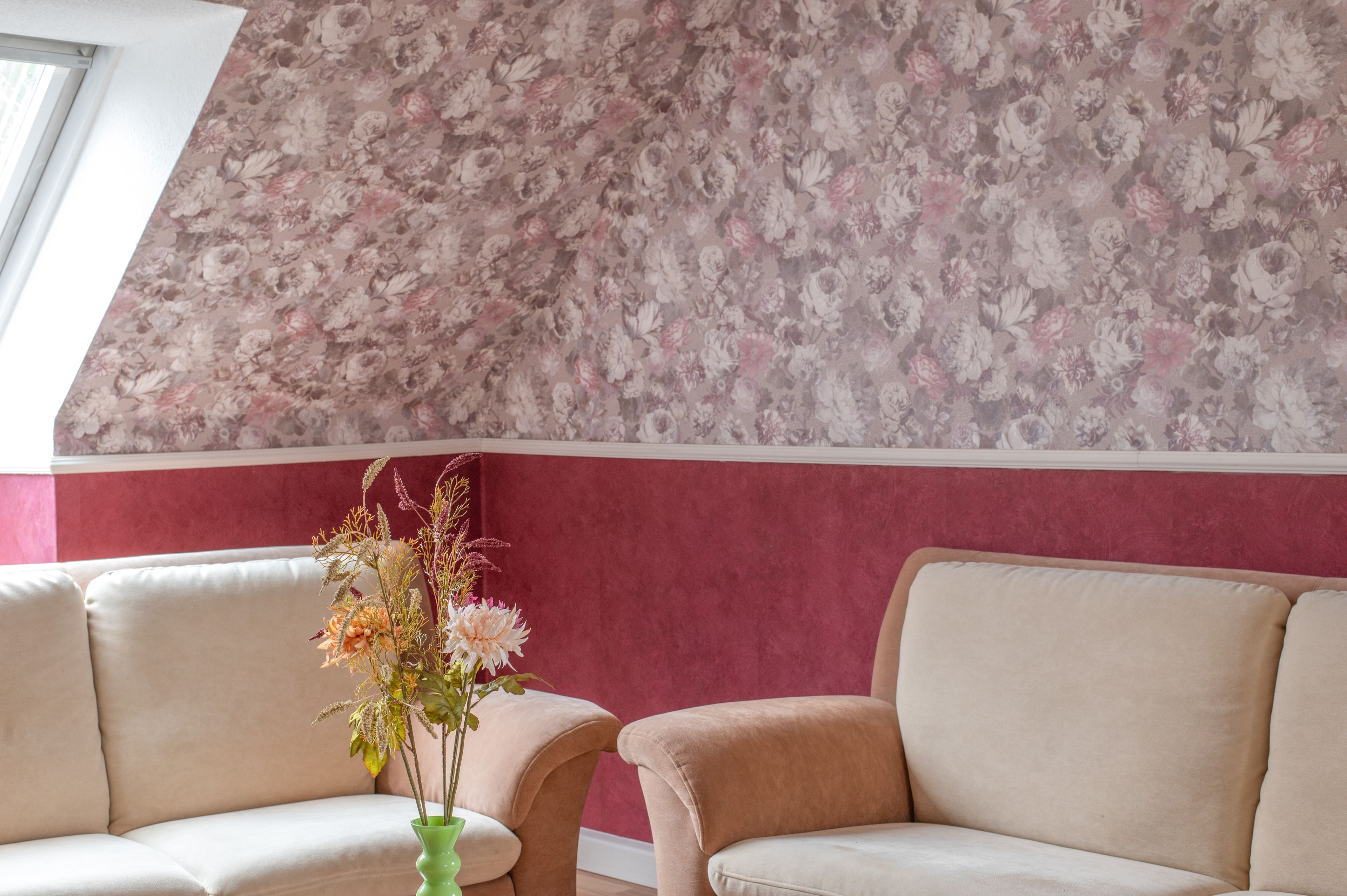 Een kamer met bloemenbehang, een groene vaas met bloemen en twee beige banken.