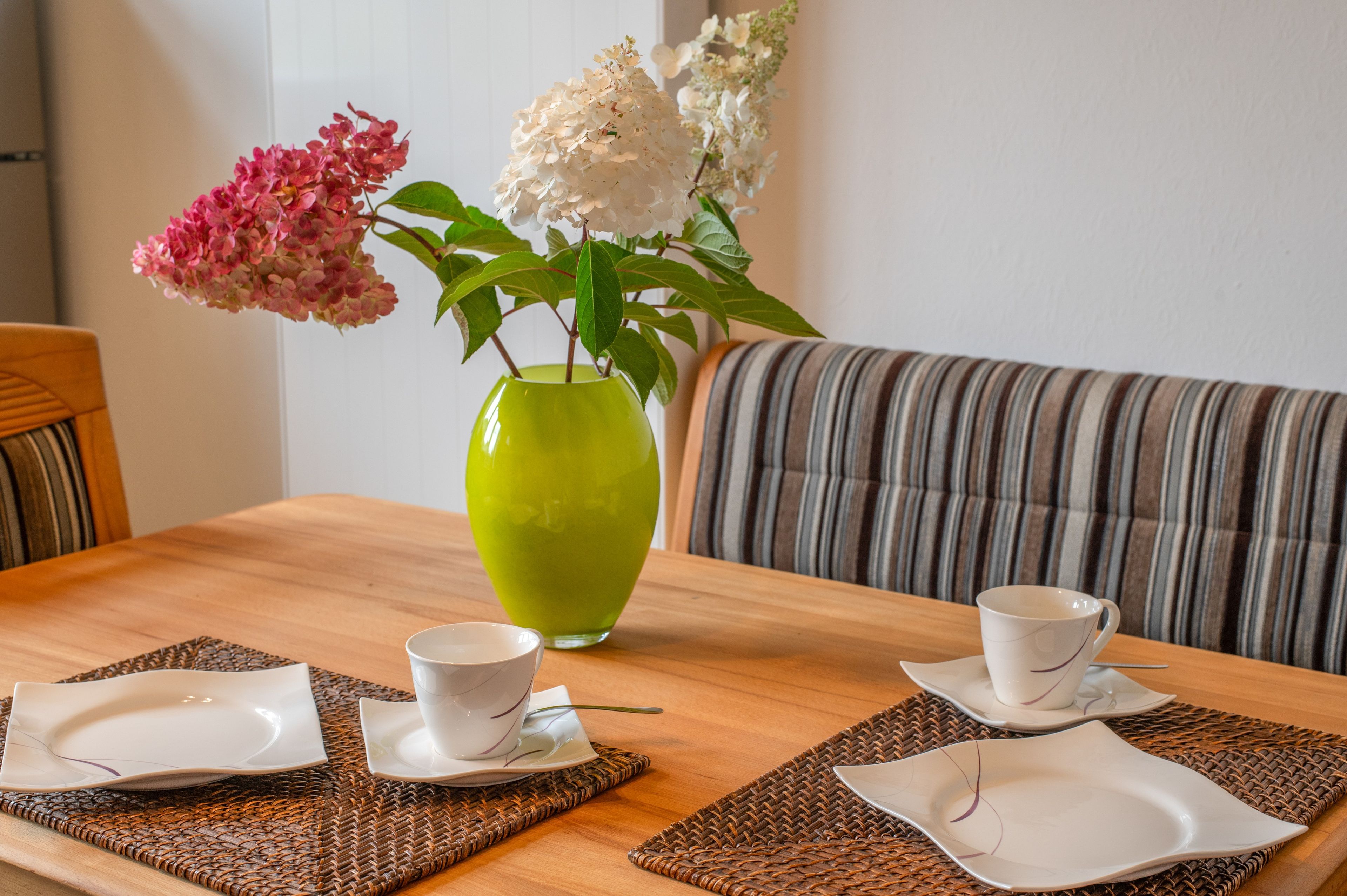 Een eettafel met twee witte kopjes en schoteltjes, een groene vaas met bloemen, en een gestreept bankstel op de achtergrond.