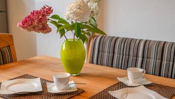 Een eettafel met twee witte kopjes en schoteltjes, een groene vaas met bloemen, en een gestreept bankstel op de achtergrond.