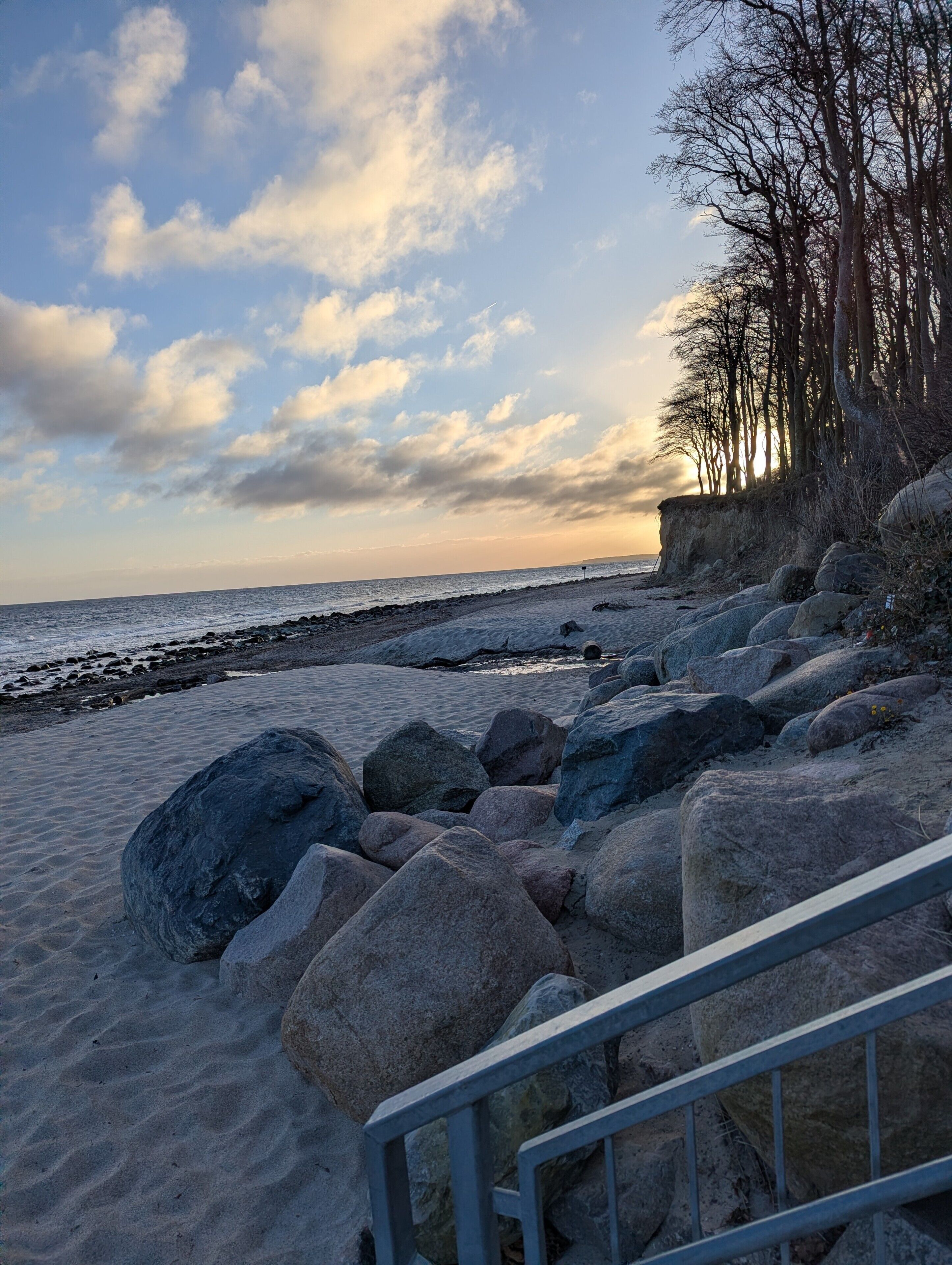 A rocky beach with a metal railing, a clear sky, and a distant view of the sea.