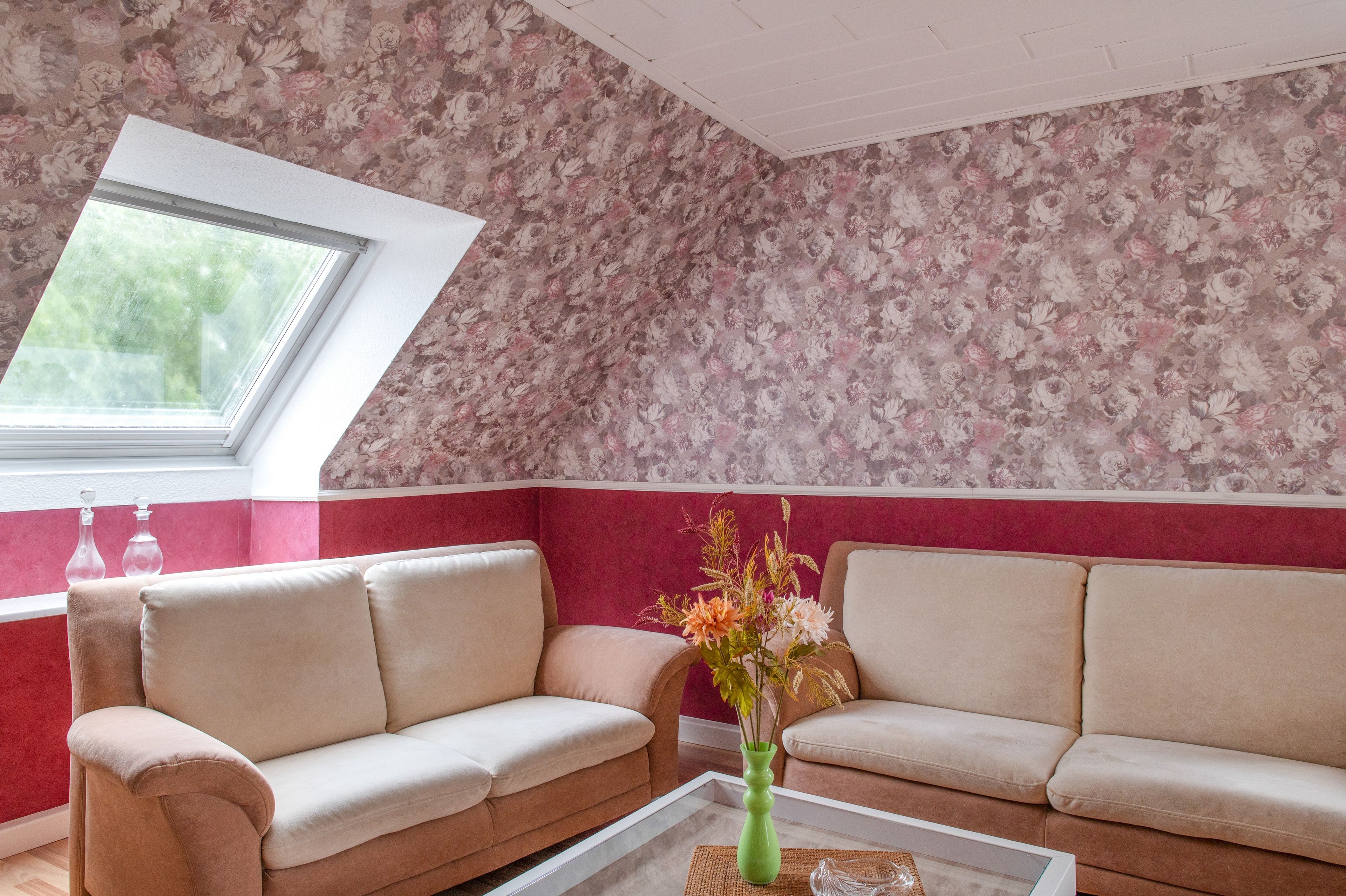 Ein Zimmer mit zwei beigefarbenen Sofas, einer grünen Vase mit Blumen, einem Fenster mit Blick auf Grünflächen und floralem Wandpapier.