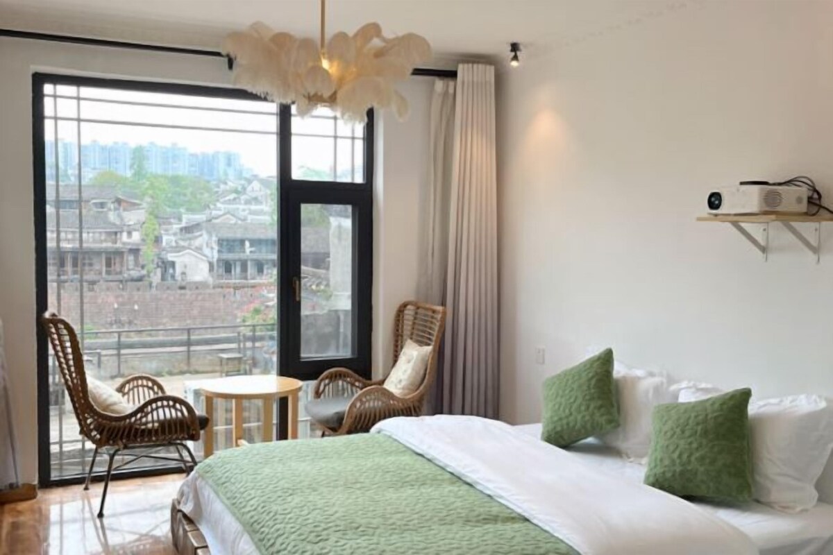 Habitación de hotel con un ventanal grande, una cama con almohadas, una silla, una mesita y una lámpara de techo.