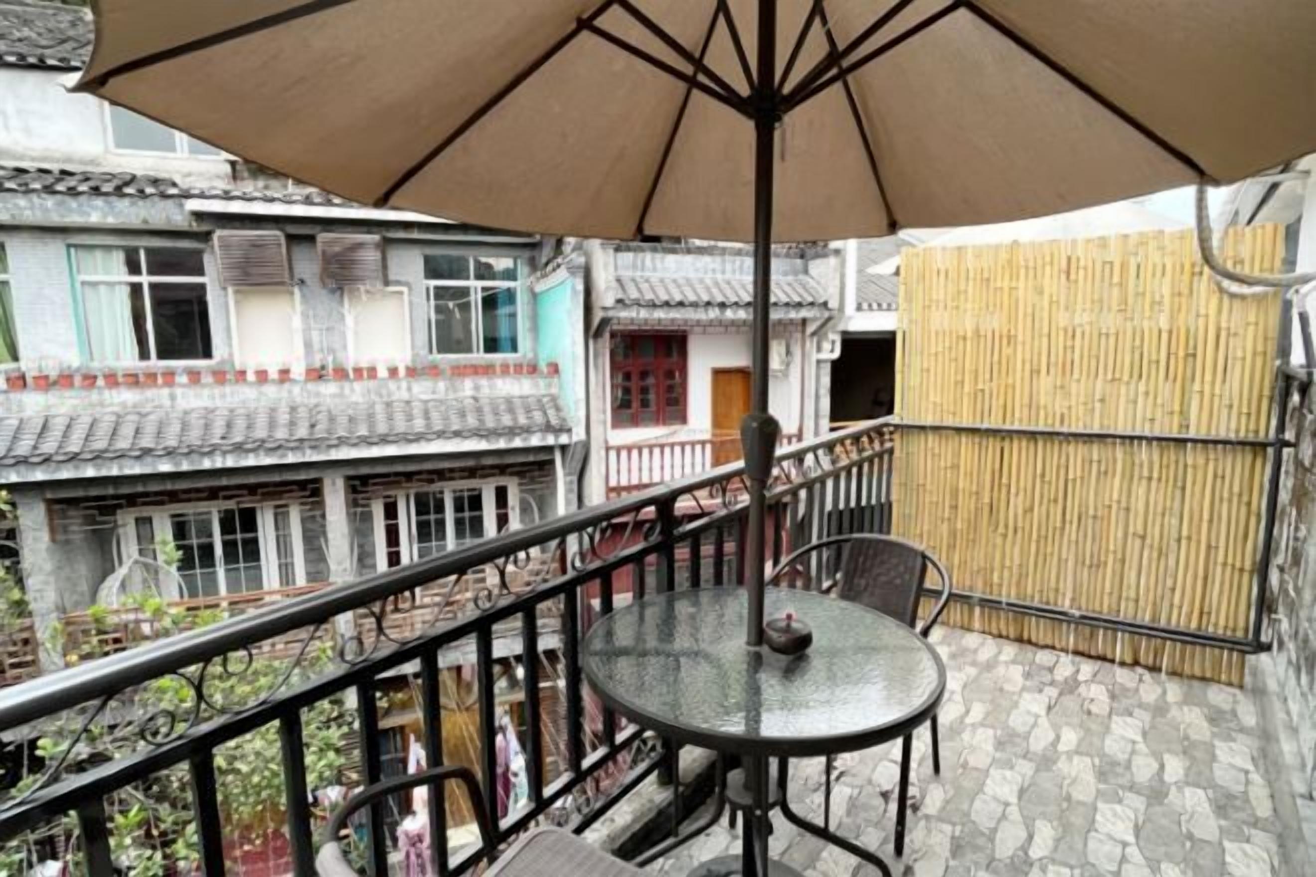 Un balcón con una mesa y una silla, un biombo de bambú y edificios tradicionales con tejas al fondo.