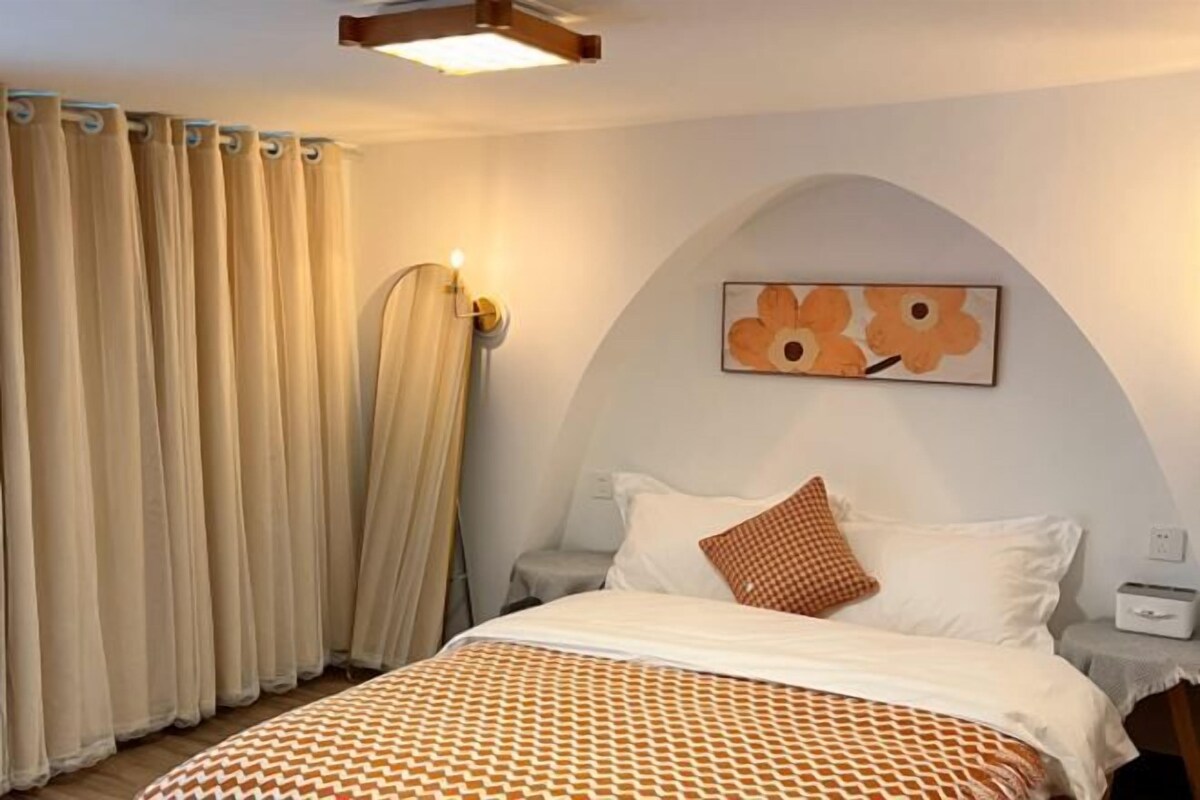 Un dormitorio con cama, cortinas, una lámpara de pared y un cuadro.