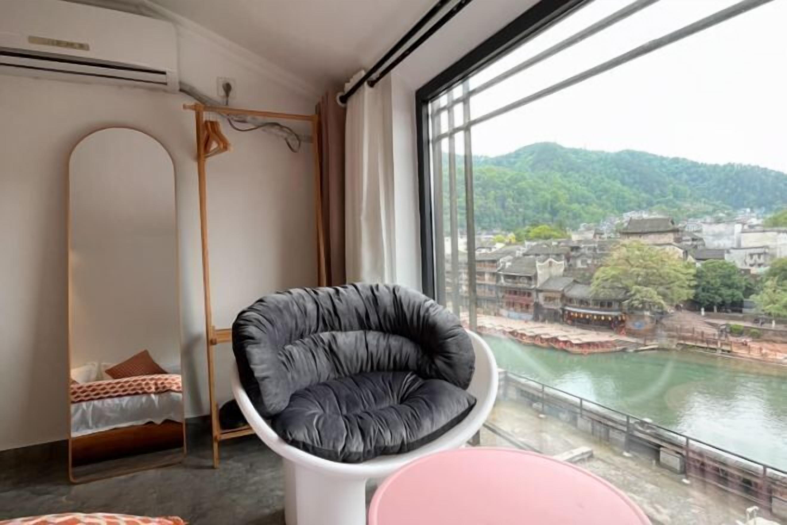 A room with a view of a river and hills, a black chair, a white table, and a bed with a mirror.