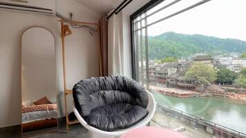 A room with a view of a river and hills, a black chair, a white table, and a bed with a mirror.