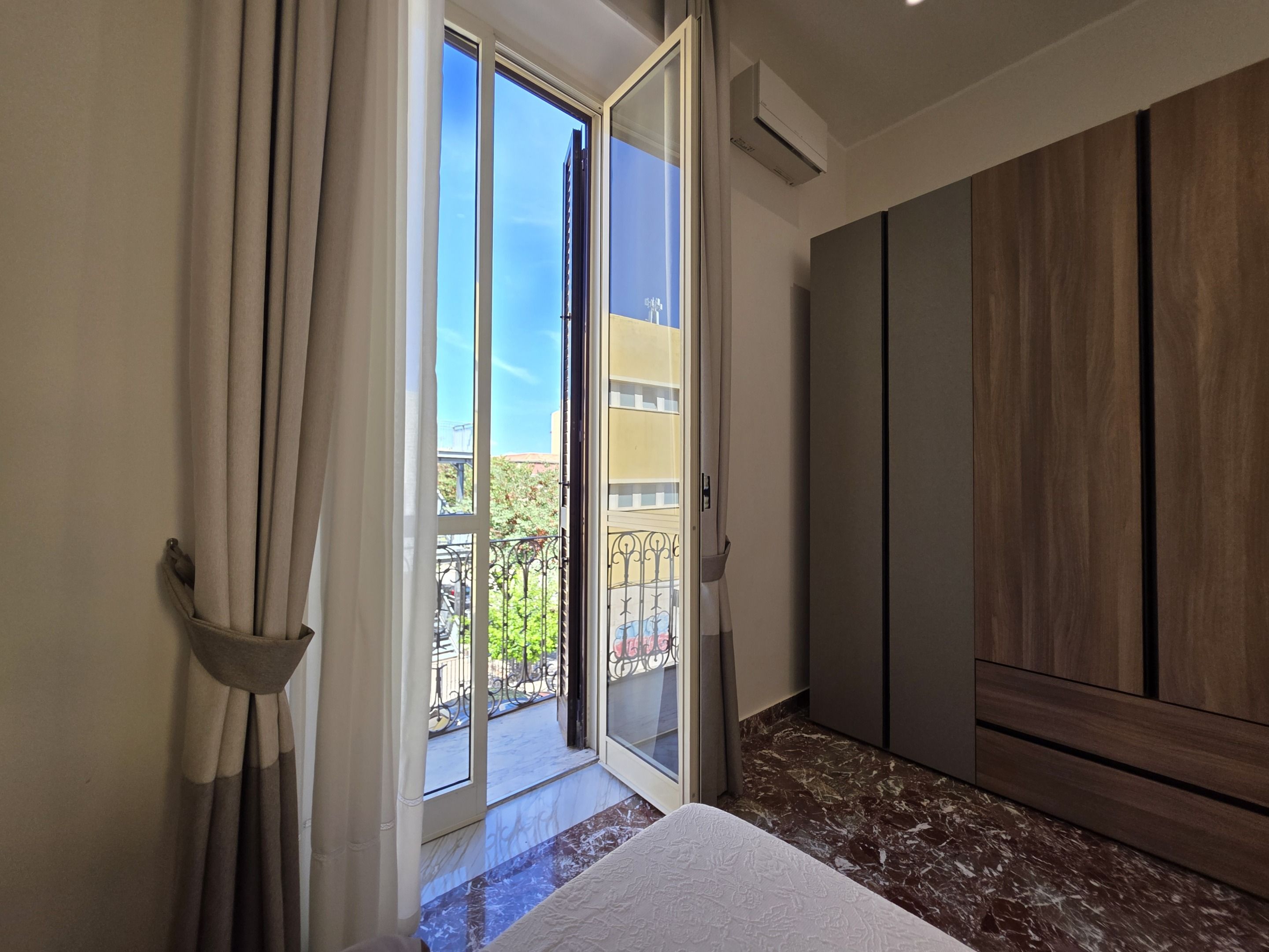 A room with a sliding glass door leading to a balcony, a bed with a white bedspread, and a wooden wardrobe.