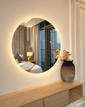A bedroom with a round mirror, a bed, a nightstand, a vase with flowers, and a view of tall buildings through the window. - Gogoto11 2BR Premium Infinity Pool & Burj View (Dubai)