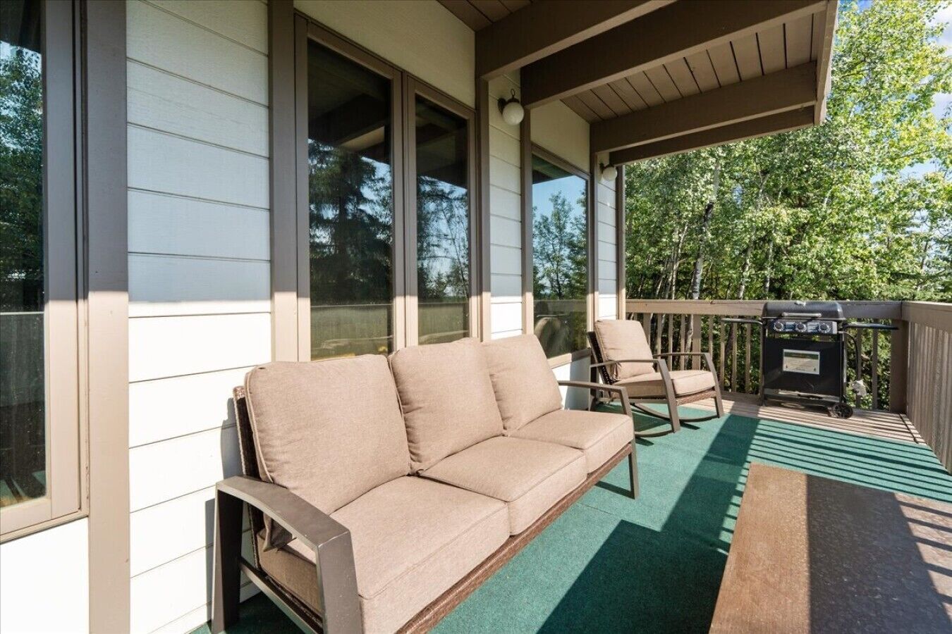 A covered porch with a beige sofa and two chairs, a grill, and a view of trees.
