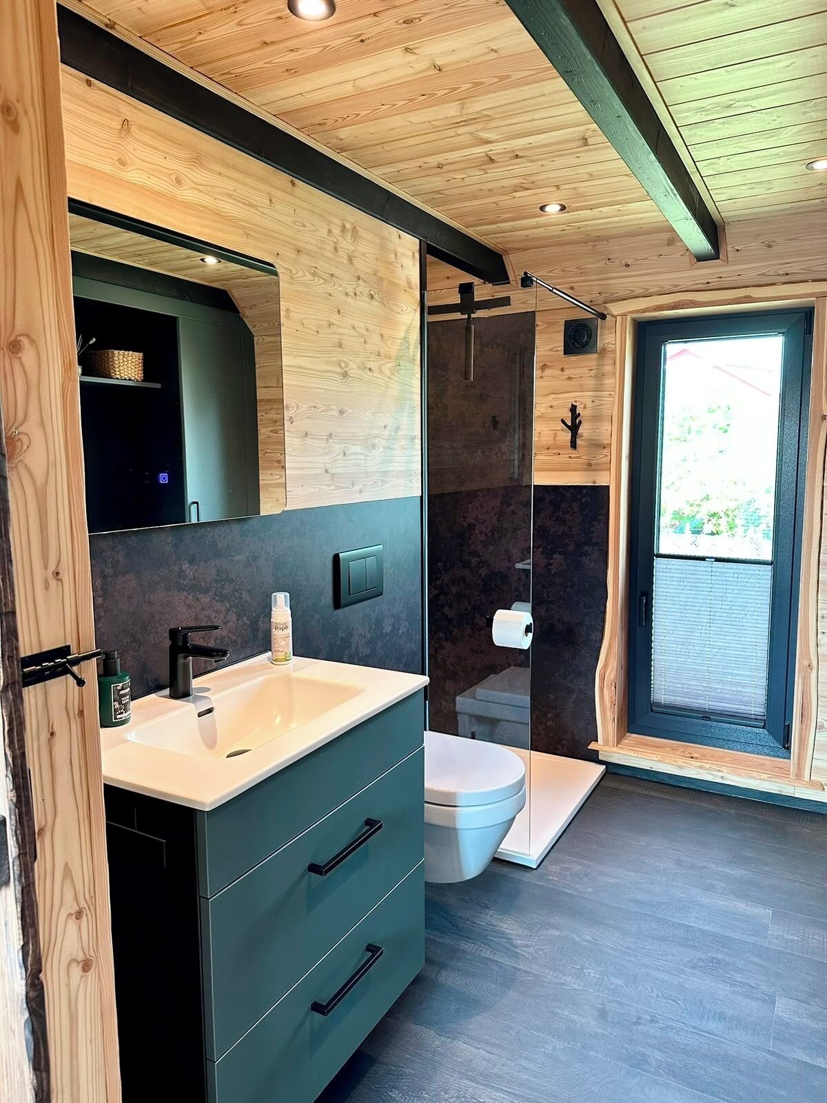 Ein modernes Badezimmer mit Holzboden, einem großen Spiegel, einem Waschbecken mit Armatur, einer Toilette und einem Duschbereich.