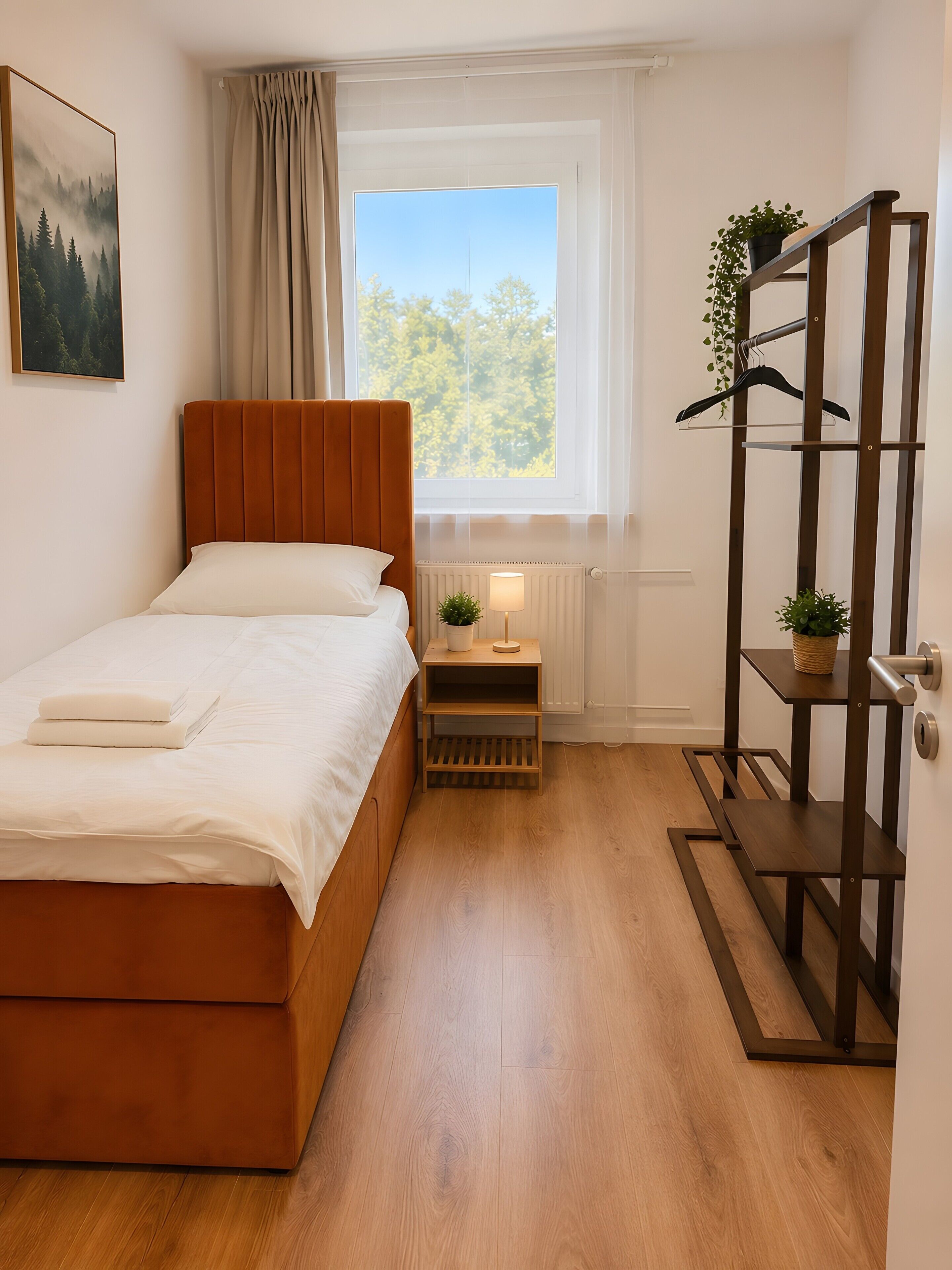 Ein Schlafzimmer mit einem Bett, einem Nachttisch, einem Fenster mit Blick auf Bäume und einem Holzboden.
