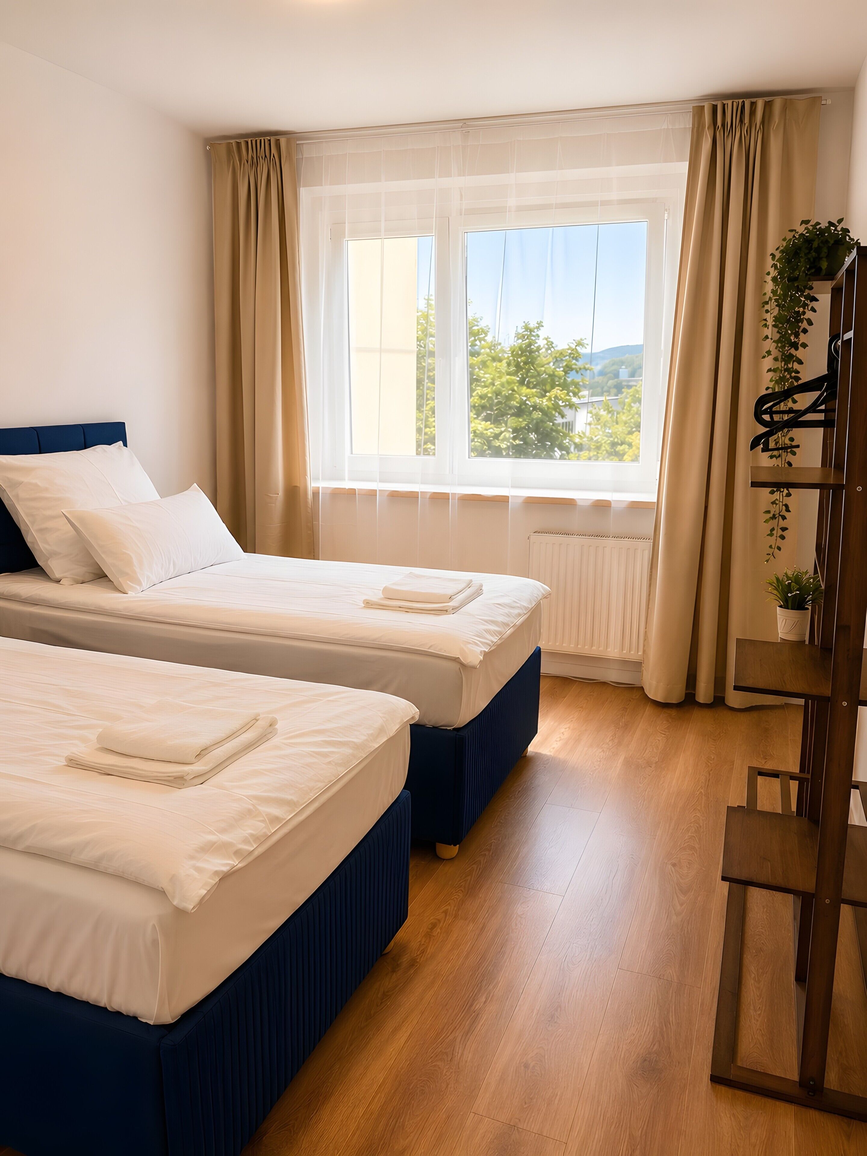 Ein Hotelzimmer mit zwei Betten, einem Heizkörper, einem mit Vorhängen versehenen Fenster und einer Pflanze auf einem Regal.