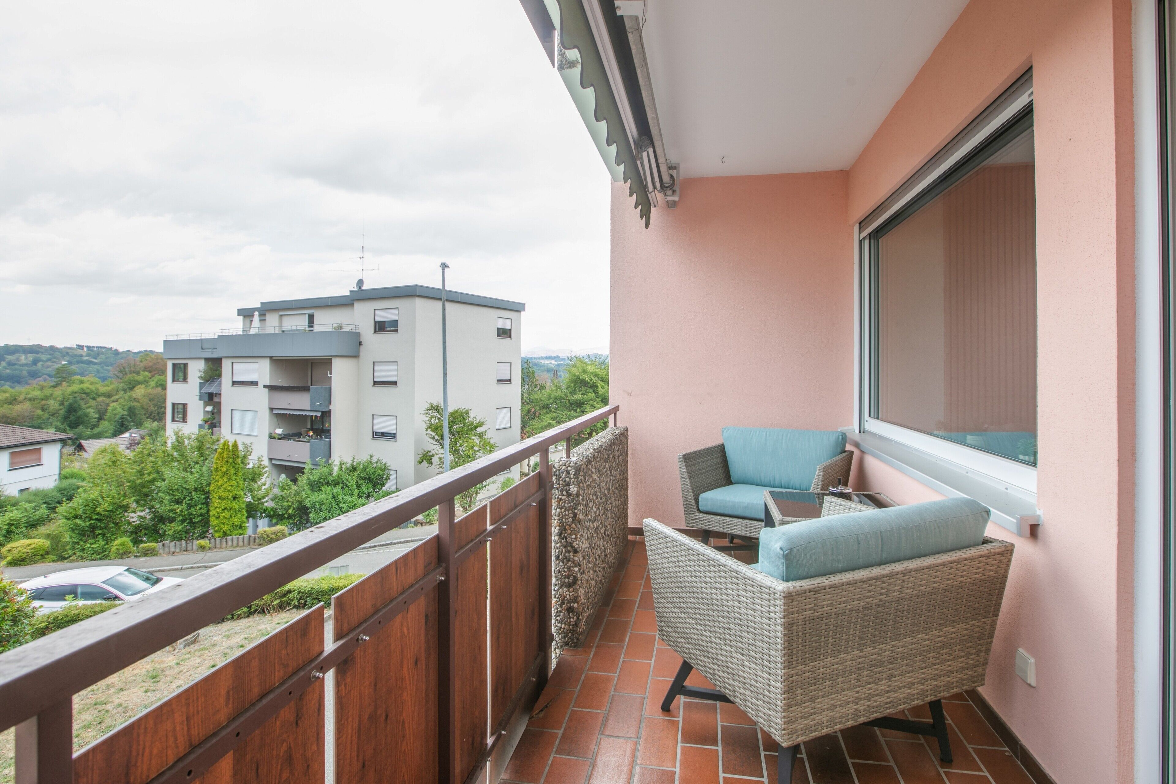 Ein Balkon mit Korbmöbeln, Blick auf ein Gebäude und ein Holzzaun.