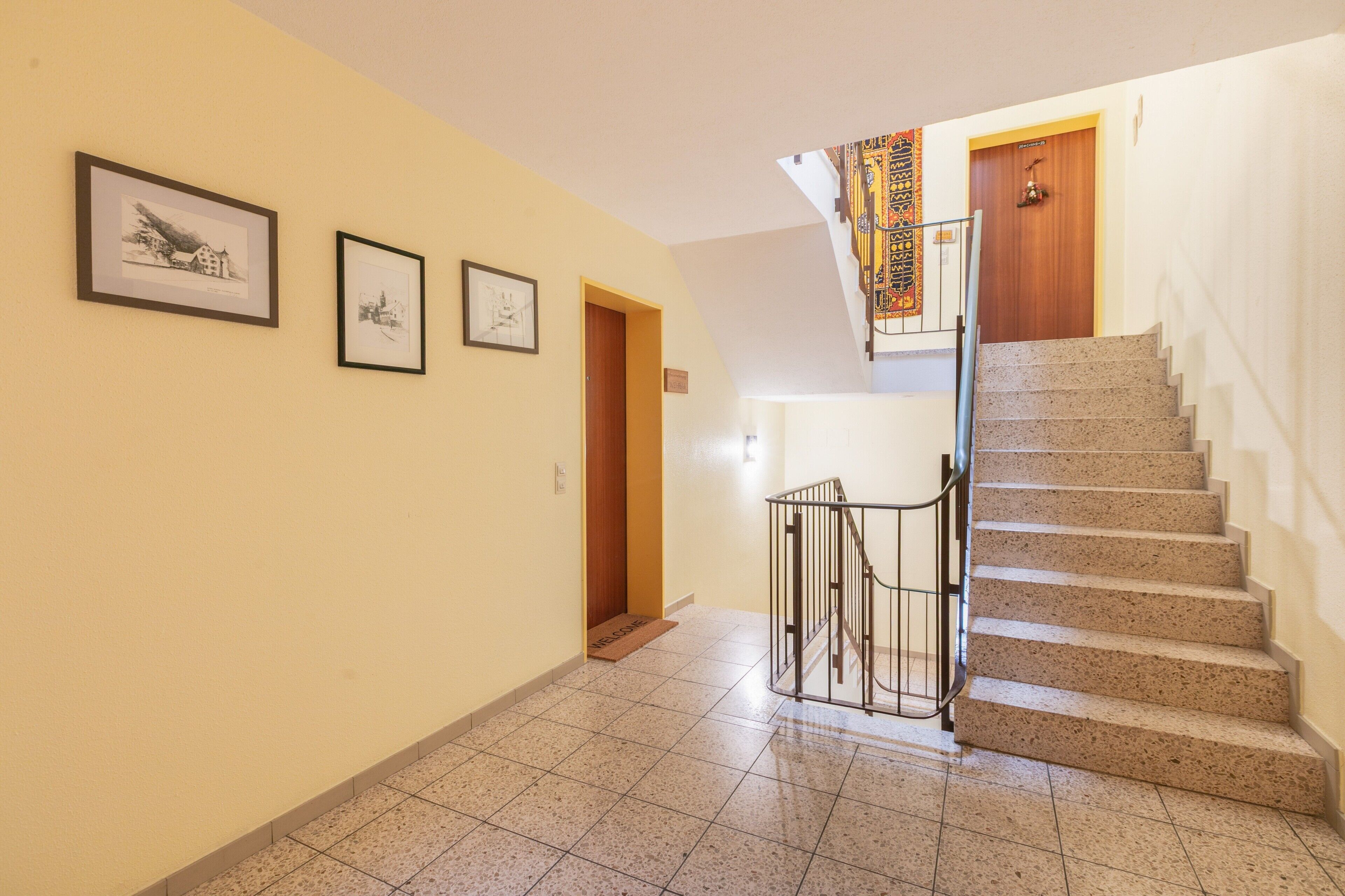 A hallway with a staircase leading to a wooden door, three framed pictures on the wall, and a welcome mat at the bottom of the stairs.