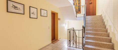 A hallway with a staircase leading to a wooden door, three framed pictures on the wall, and a welcome mat at the bottom of the stairs.