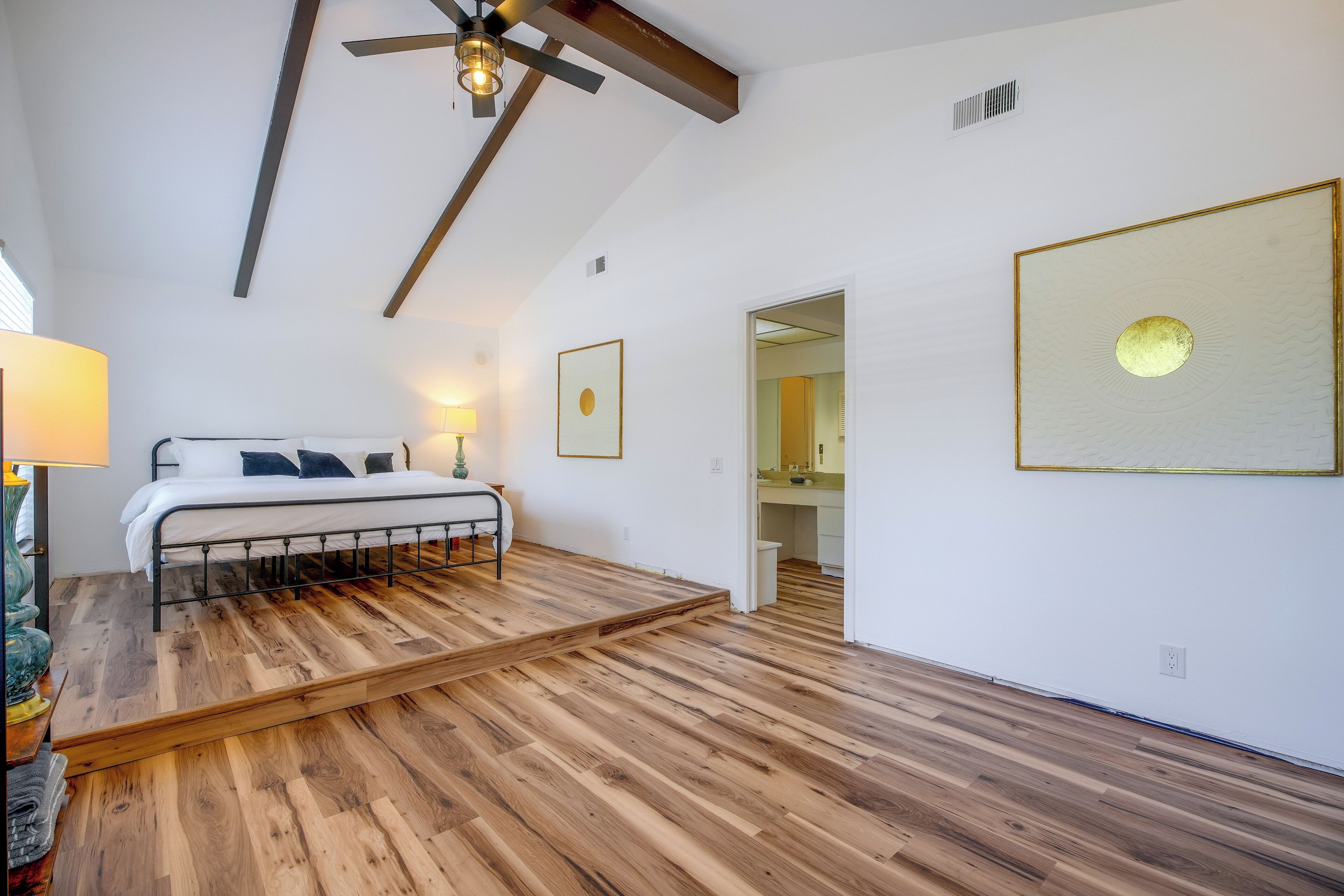 Ein Schlafzimmer mit einem Bett, einem Deckenventilator, Holzboden, einer Wandleuchte und einem gerahmten Bild.