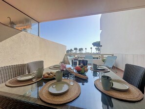 An outdoor dining area with a glass table, wicker chairs, and a view of palm trees and a building in the background. - Ulysse Port de Plage by Interhome (Port Camargue)