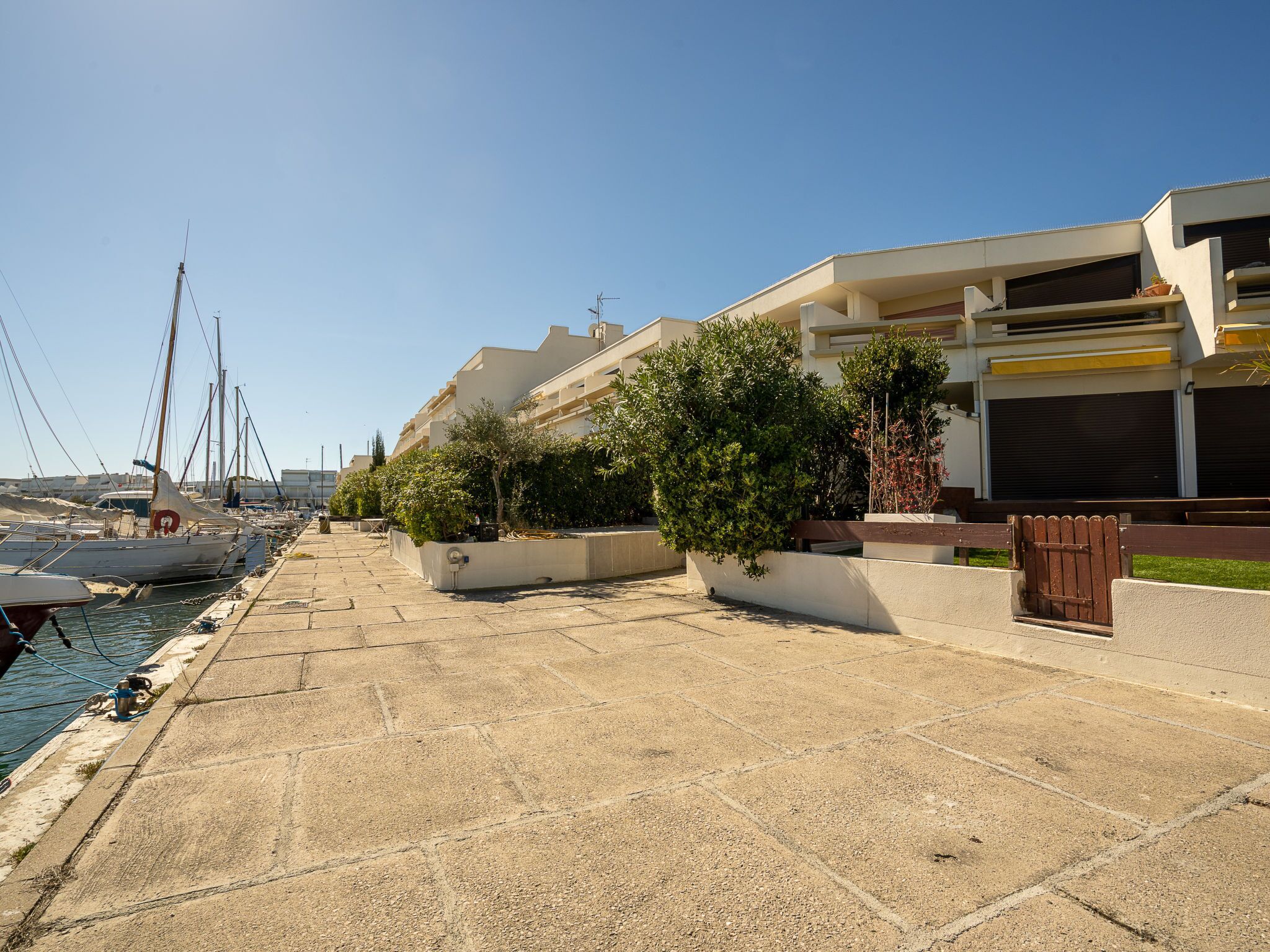 A marina with boats docked, a paved walkway, and modern buildings with balconies.