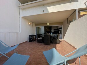 An outdoor patio area with a dining table and chairs, a small kitchen visible through an open door, and a trash bin. - Ulysse Port de Plage by Interhome (Port Camargue)