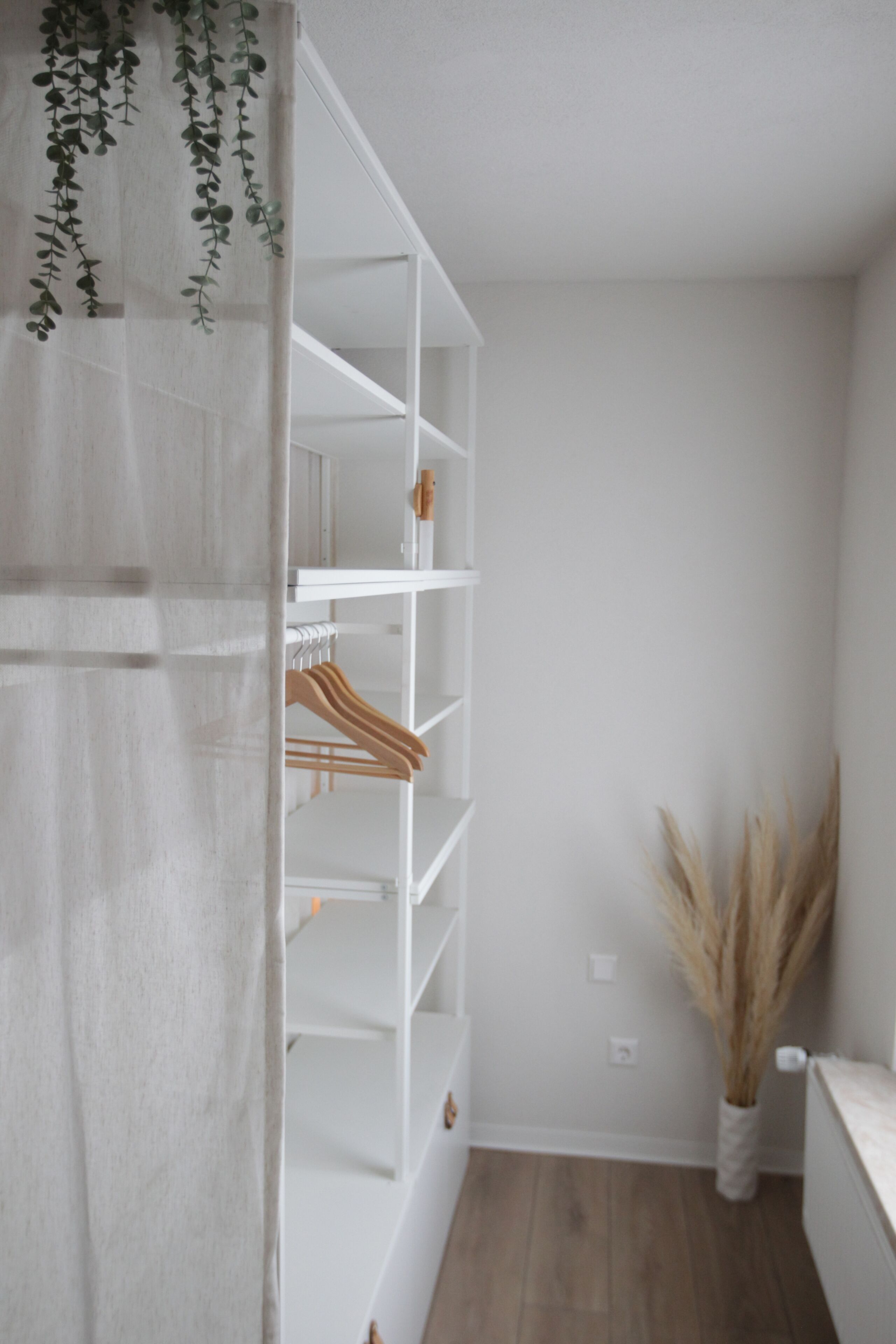 Un armario blanco con varios estantes y una barra para colgar, suelo de madera y un florero con plantas secas sujeto a la pared.