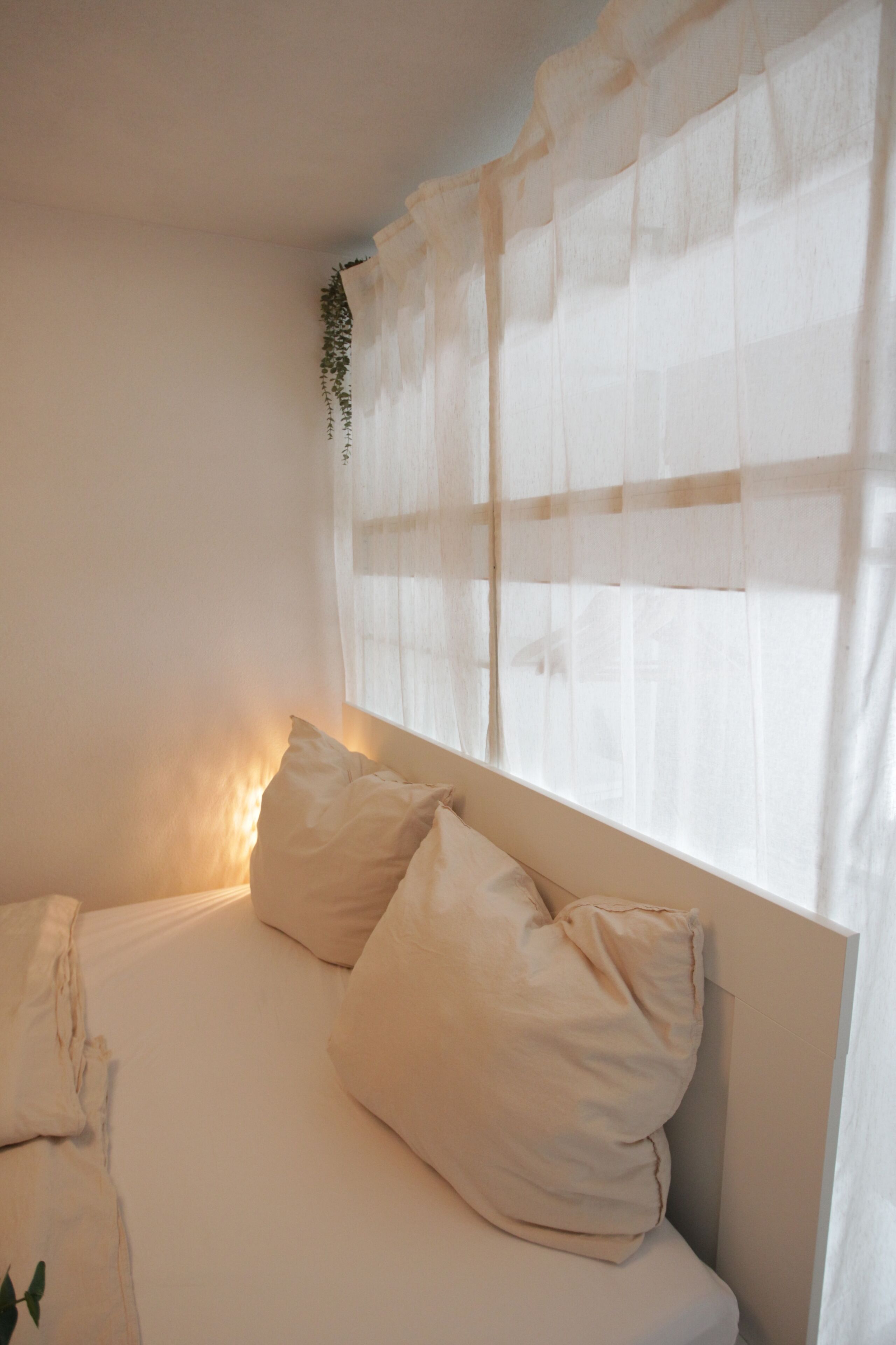 Ein Schlafzimmer mit einem Bett, Kissen, einem Fenster mit durchsichtigen Vorhängen und einer Wandpflanze.