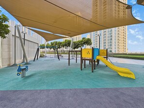 A playground with a slide and swings, surrounded by a shaded area and tall buildings in the background. - Bloomfields 1BR in Creekside 18 (Dubai)