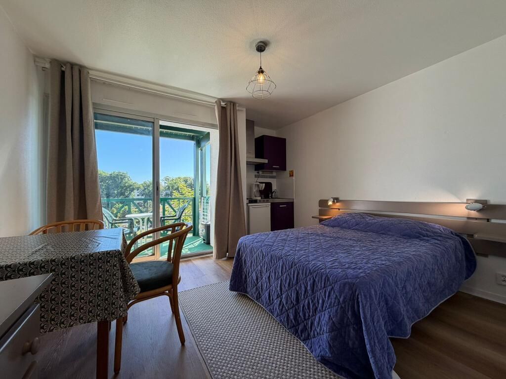 A bedroom with a bed, a desk, and a chair. There is a balcony with a view of trees and a building.