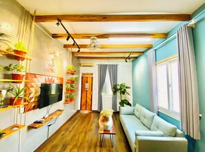 Ein Wohnzimmer mit Holzbalkendecke, einem Flachbildfernseher, einer Couch, einem Couchtisch und verschiedenen Kübelpflanzen.