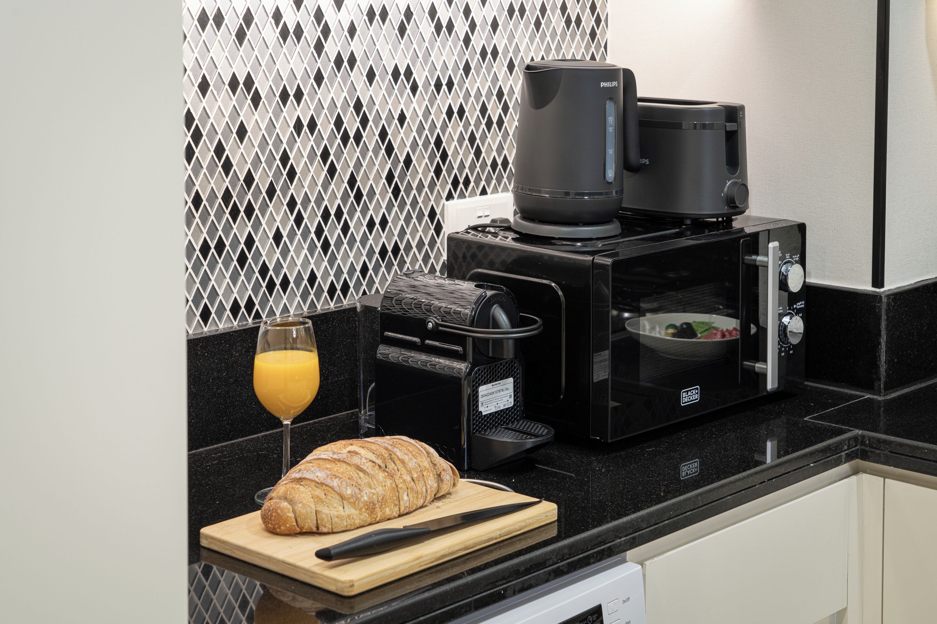 Una cocina con cafetera, microondas, tostadora, un vaso de jugo de naranja y una barra de pan sobre la encimera.