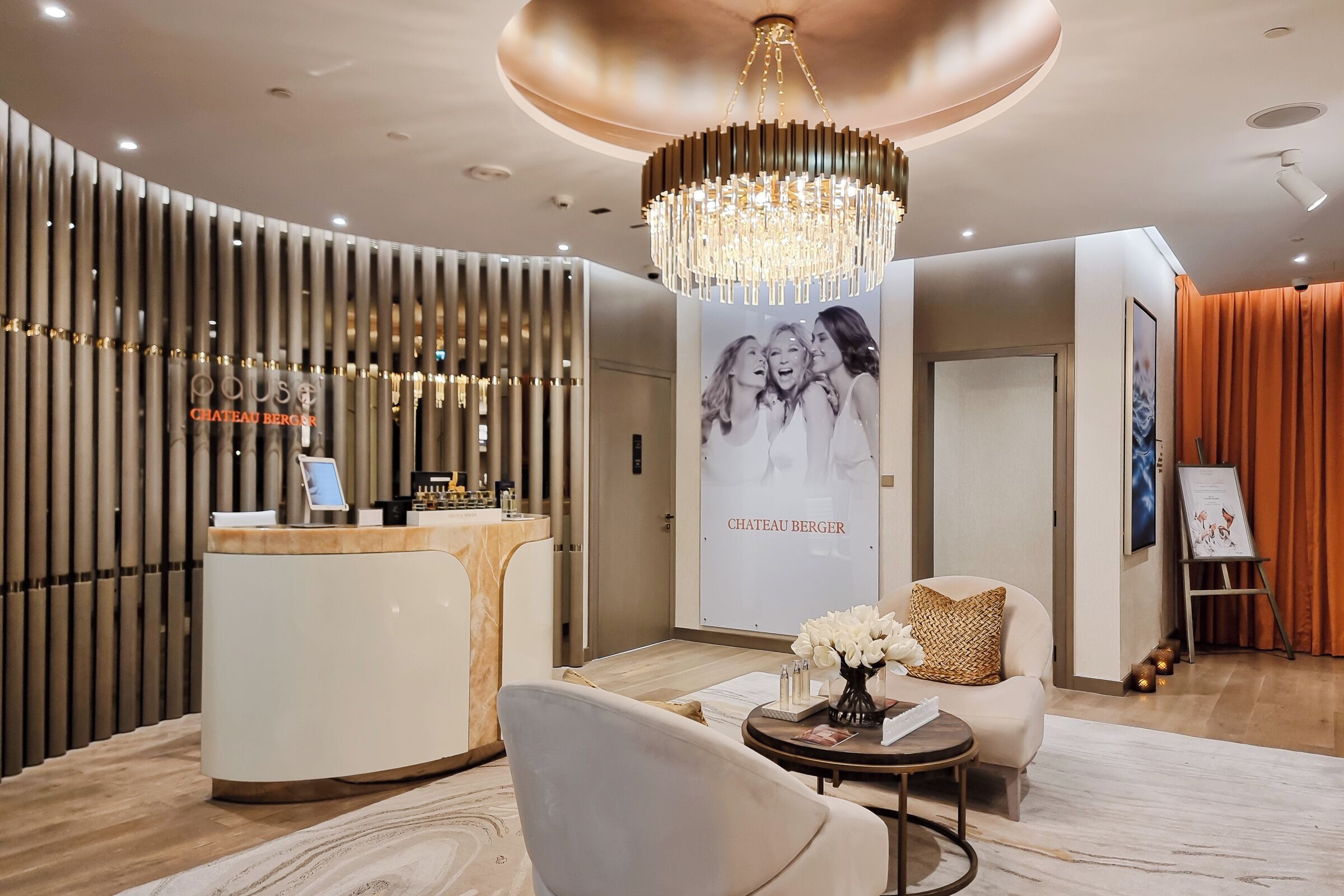 A modern reception area with a reception desk, a chandelier, a seating area with a sofa and armchair, and a wall with a poster.