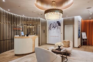 A modern reception area with a reception desk, a chandelier, a seating area with a sofa and armchair, and a wall with a poster. - Stylish 2BR in Paramount Infinity Pool   (Dubai)