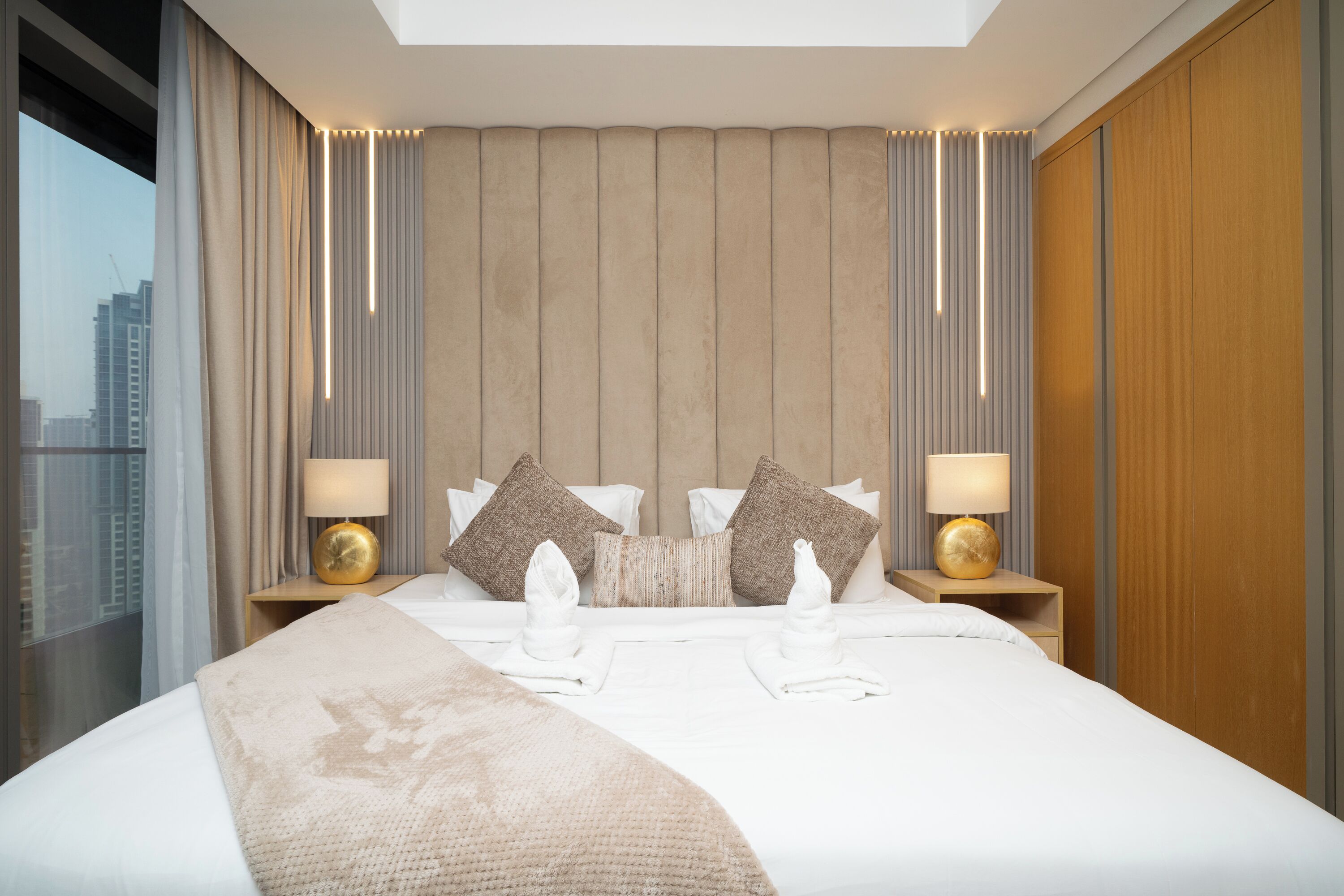 A hotel room with a large bed, two bedside tables with lamps, a headboard with vertical lines, and a view of the city through the window.