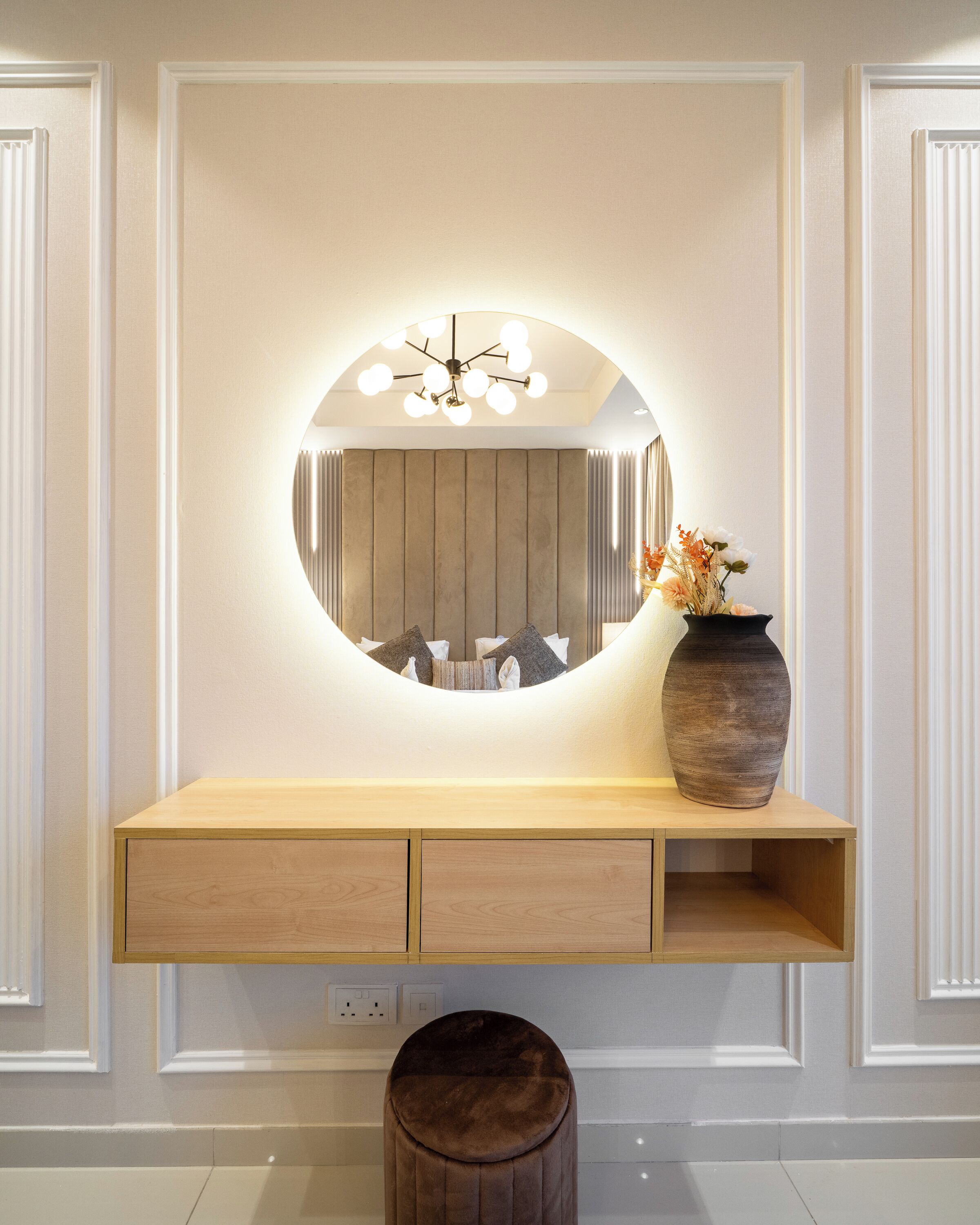 A vanity with a round mirror, a vase with flowers, and a wooden stool.