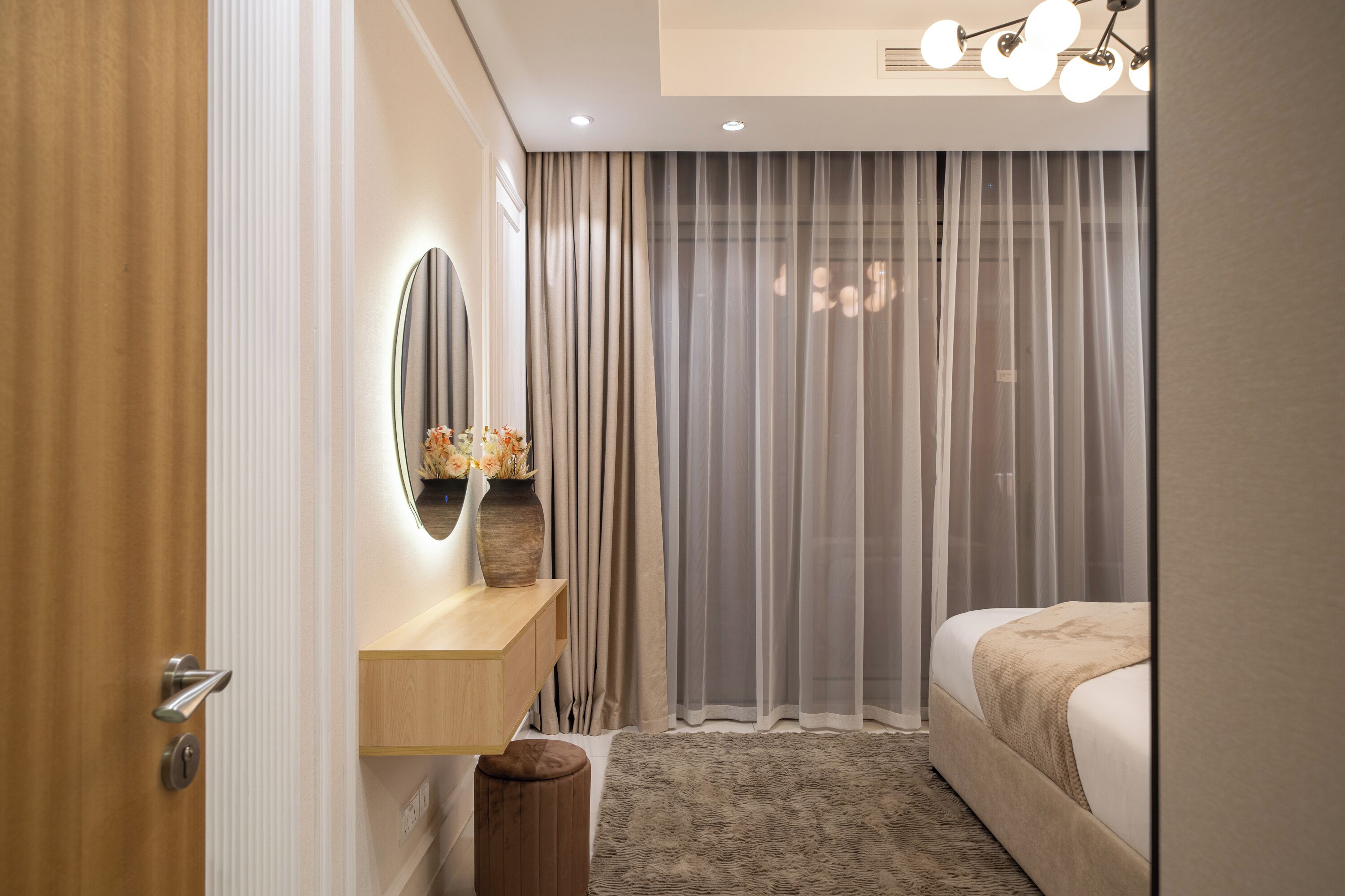 A modern bedroom with a wooden vanity, a round mirror, a bedside table, a bed with a beige blanket, and a large window with sheer curtains.