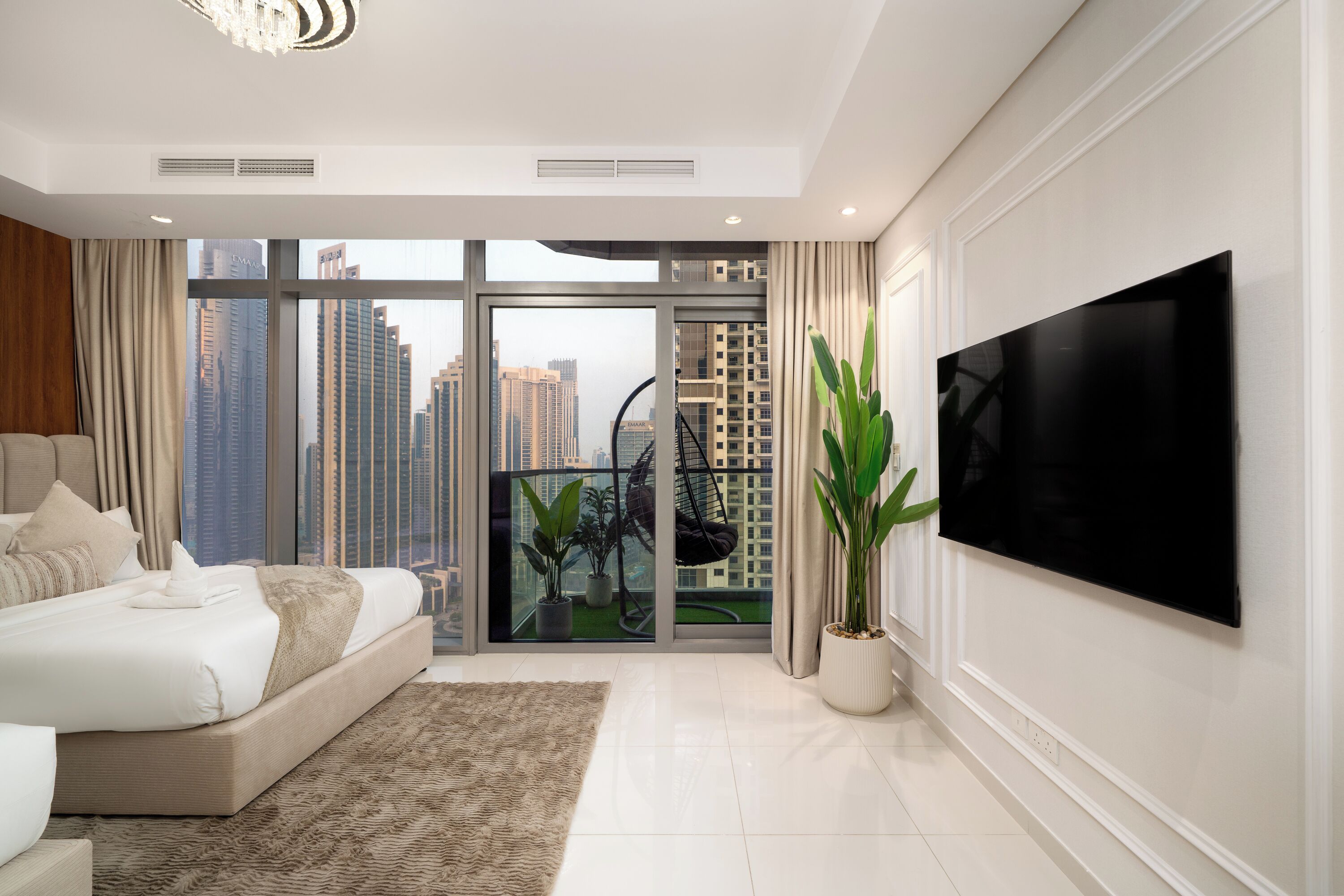 A modern bedroom with a large bed, a TV, and a balcony with a view of high-rise buildings.