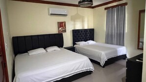 Habitación de hotel con dos camas, ventilador de techo, equipo de aire acondicionado, un cuadro en la pared y una ventana con cortinas.