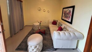 Una sala de estar con un sofá blanco, un otoman redondo, una mesa de centro de vidrio y un cuadro en la pared.