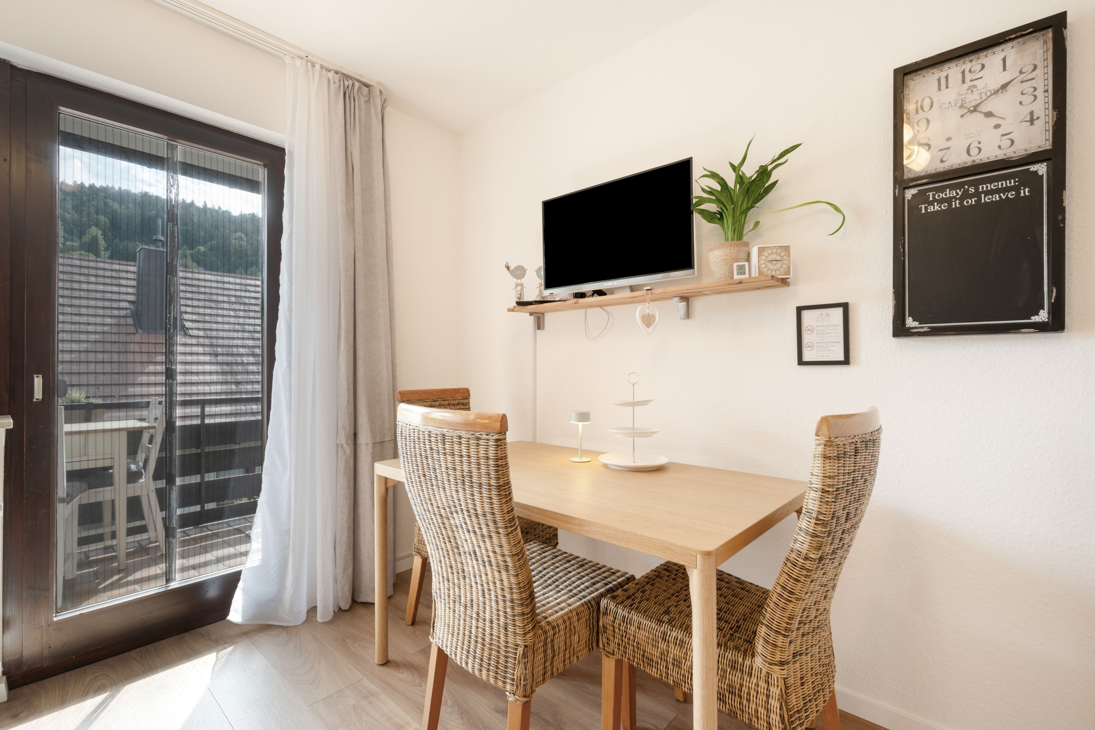 A dining area with a wooden table and wicker chairs, a mounted TV, a potted plant, and a wall clock.