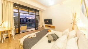 Habitación de hotel con una cama, un escritorio con silla, un televisor y un ventanal con vistas a edificios.