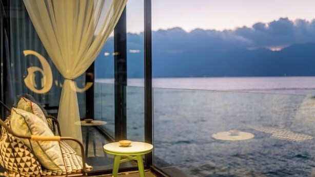 Una habitación con vista al agua, un separador de vidrio, una butaca acolchada y una pequeña mesa redonda.