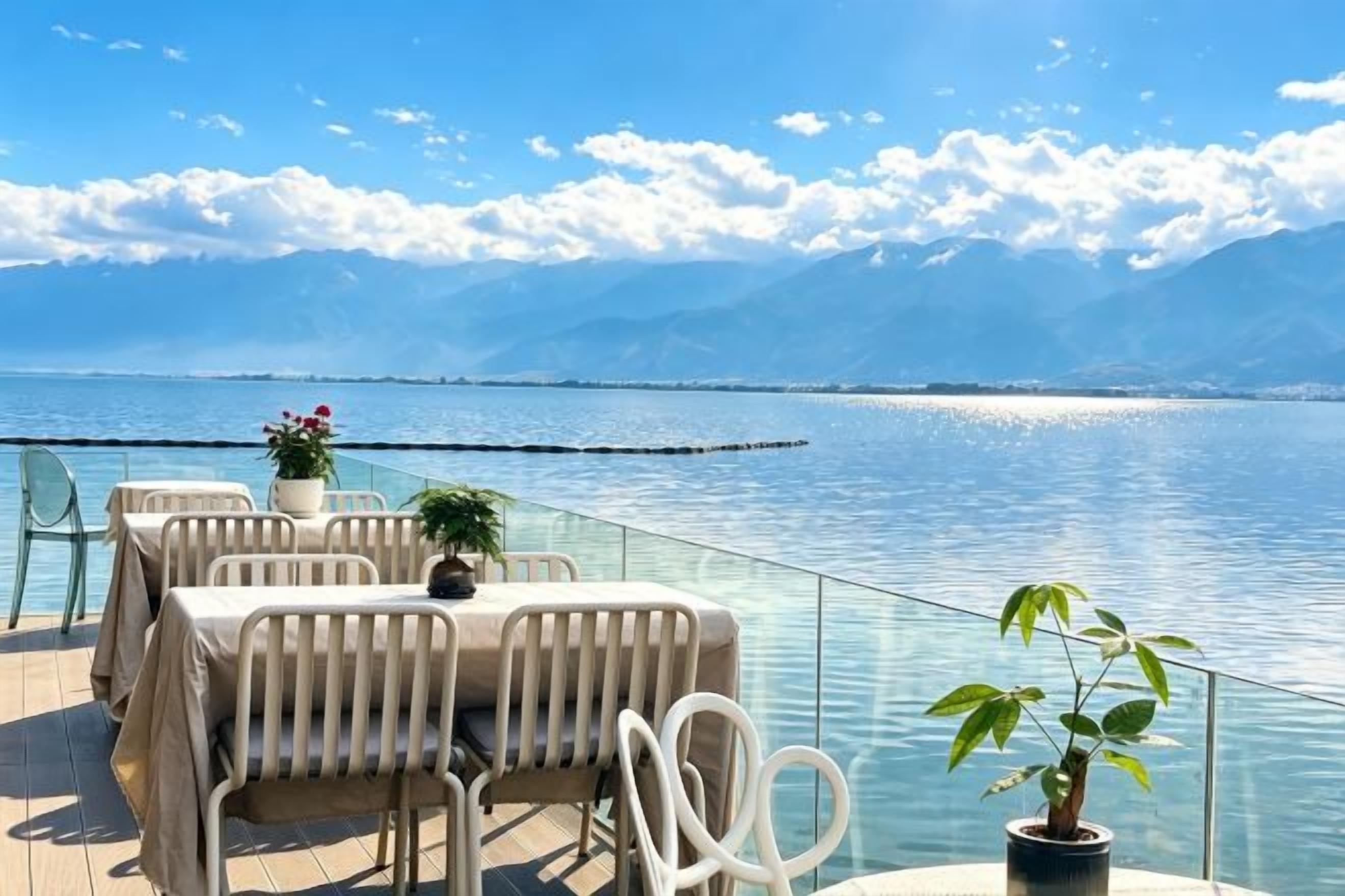 Un área de comedor al aire libre con mesas y sillas, con vista a un cuerpo de agua y montañas.