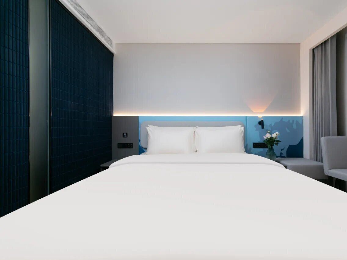A hotel room with a large bed, a nightstand, a chair, and a wall-mounted lamp.