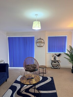 Ein Wohnzimmer mit blauem Sofa, einem runden Couchtisch mit Fernbedienung, einem Korbstuhl, einem kleinen Beistelltisch mit Dekoration und einer Topfpflanze.