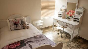 A bedroom with a bed, a dresser, a vanity with a mirror, and a window with blinds.